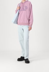 Fioletowa bluza z kapturem z logo 'GAP', jasnoniebieskie dżinsy i czarne buty na platformie. Trzyma jasnoniebieską torbę na ramię. Prosty i casualowy design.