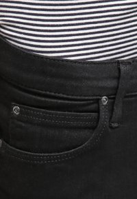 Jeans in denim nero con una vista ravvicinata della vita, dotati di un bottone argentato e di una tasca anteriore. Un top a righe è parzialmente visibile sopra.