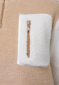 Bézs és fehér polár anyag világosbarna, függőleges cipzárral a fehér folton, puha és texturált anyagot mutatva.