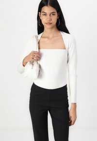 Valkoinen pitkähihainen bodysuit, jossa on neliön muotoinen pääntie, yhdistetty mustiin korkeavyötäröisiin housuihin. Malli pitää pientä valkoista käsilaukkua.