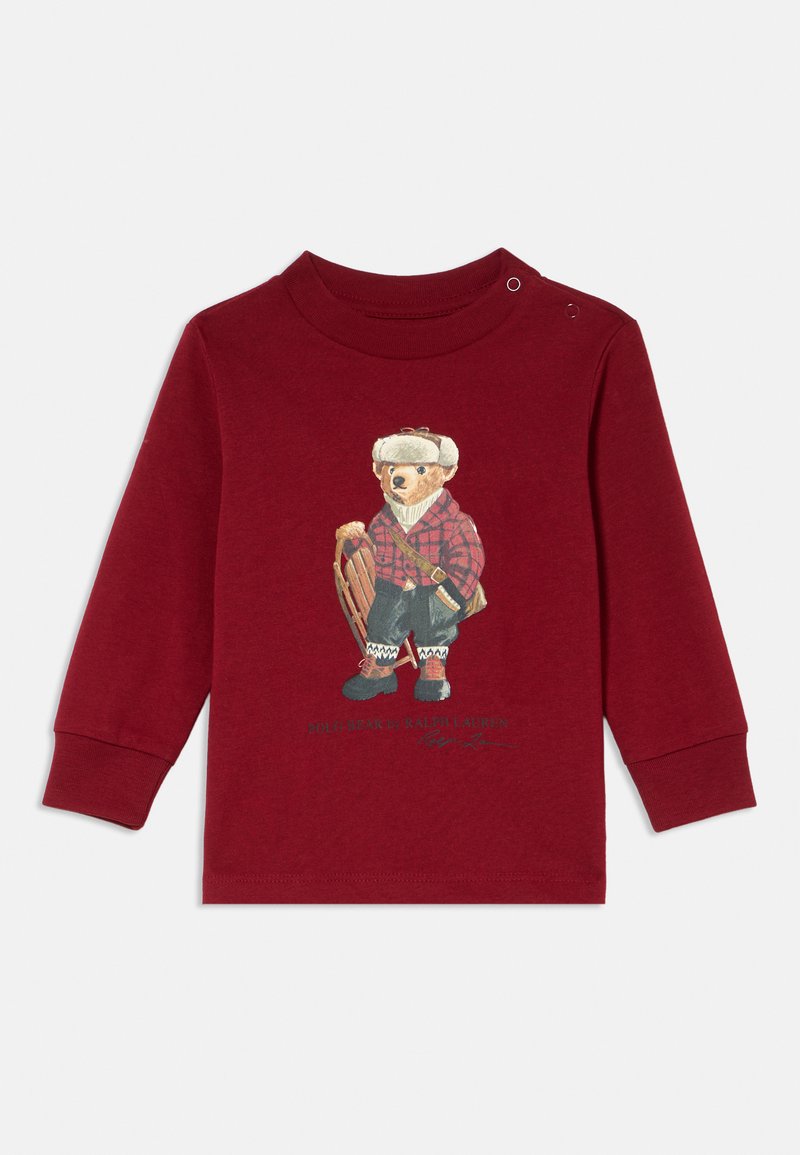 T-shirt vermelho de manga longa com uma ilustração de um urso em uma camisa xadrez, segurando um trenó. Inclui um botão circular na gola.