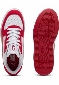 Chaussures de sport blanches et rouges avec une tige synthétique lisse, des accents texturés, des perforations sur la pointe et une semelle extérieure en caoutchouc présentant un motif de crampons.