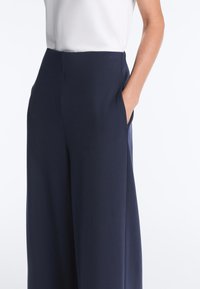 Kvinde iført marineblå, højttaljede bukser med vide ben, med hånden i lommen og en hvid, ærmeløs top, der er stoppet ind i bukserne.