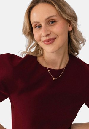 Junge Frau mit blondem Haar, die ein burgunderfarbenes Oberteil und eine zarte goldene Halskette trägt, lächelt sanft vor einem schlichten, hellen Hintergrund.