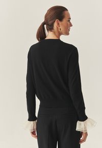 Crna sweatervesta s dugim rukavima koja ima prozirne, nabranih rukave. Dizajn je prilagođen s okruglim izrezom i blago skraćenom dužinom.