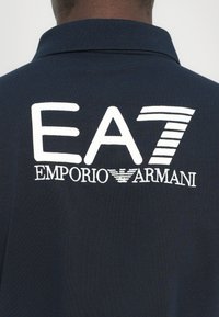 Polo de cor azul marinho escuro com um logo bordado branco "EA7 EMPORIO ARMANI" nas costas, com colarinho clássico e textura suave.