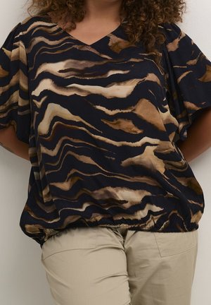 Kobieta w luźnym czarnym topie z brązowymi i beżowymi abstrakcyjnymi paskami przypominającymi tygrysa oraz beżowych spodniach, stojąca na tle jednolitego jasnego tła.