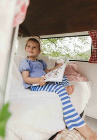 Kind dat op een witte bank zit, gekleed in een blauwe top met pofmouwen en blauw-wit gestreepte leggings, en een boek leest. Zachte texturen en subtiele patronen zijn zichtbaar.