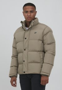 Beige puffer jacket with a quilted design, round collar, and black buttons. Features a small logo on the chest and a zippered closure.