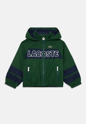Grüne Kapuzenjacke mit navyfarbenen Akzenten, featuring "LACOSTE" Text, elastischen Bündchen und Reißverschlusstaschen. Logo auf der oberen linken Seite.