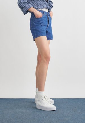 Persona con shorts de mezclilla azul, zapatillas blancas de caña alta con calcetines blancos y una camisa azul de cuadros con las manos en los bolsillos, de pie sobre una alfombra azul.