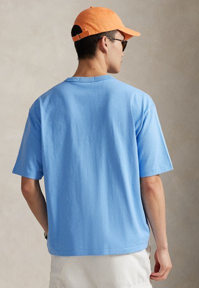 Ανοιχτό μπλε βαμβακερό μπλουζάκι με κοντά μανίκια και χαλαρή εφαρμογή, με στρογγυλή λαιμόκοψη. Φοράει πορτοκαλί καπέλο και γυαλιά ηλίου.