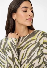 Frau mit langen, dunklen Haaren, die silberne Creolen und Ringe trägt, gekleidet in einem gemusterten Pullover mit grauen und gelben abstrakten Streifen.