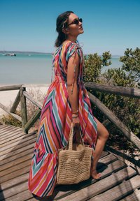 Abito maxi a righe colorato con spalline regolabili, in vivaci tonalità di rosa, blu e giallo, abbinato a una borsa di paglia beige.