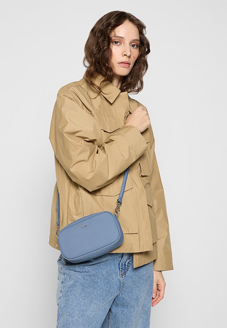 Sac en bandoulière en cuir bleu clair avec une finition texturée, de forme ovale et doté d'une sangle réglable, porté sur une veste beige et un pantalon en denim.