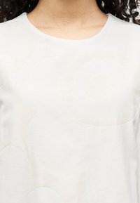 Λευκό τοπ με απαλό, υφασμάτινο λουλουδένιο μοτίβο, στρογγυλή λαιμόκοψη και απαλό ύφασμα. Χωρίς ορατά μεταλλικά στοιχεία ή διακοσμητικά.