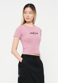 Ružičasta kratka majica s kratkim rukavima, koja ima crni izvezeni logo "Santa Cruz". Tkanina izgleda glatko i pristaje.