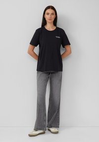 Czarny t-shirt z bawełny, krótki rękaw, okrągły dekolt, z białym napisem na klatce piersiowej, zestawiony z szarymi spodniami dzwonami i beżowymi sneakersami.