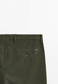 Oliivinvihreät, kapealahkeiset housut. Ne ovat litteät edestä, niissä on vyölenkit, takatasku nappikiinnityksellä ja metallinen logolaatta.