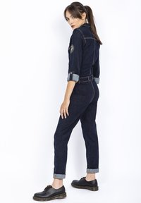 Denimowy kombinezon w ciemnoniebieskim kolorze z podwiniętymi rękawami, wcięciem w talii i kieszeniami bocznymi; zestawiony z czarnymi butami z żółtym przeszyciem.