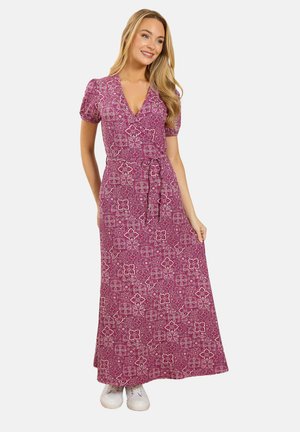 Vrouw die een maxi-jurk met patroon draagt, roze wikkeljurk met korte mouwen, een gestrikte taille en witte sneakers, glimlachend en staand tegen een witte achtergrond.