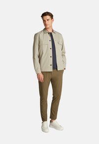 Giacca button-up grigia chiara con due tasche sul petto, abbinata a una camicia blu navy e pantaloni verde oliva, completata con sneaker bianche.