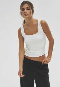 Weißes, eng anliegendes Tanktop mit quadratischem Ausschnitt, getragen mit schwarzen Jogginghosen mit Kordelzug. Der Stoff wirkt glatt und elastisch.