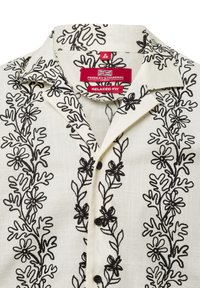 Camicia bianca con ricami floreali neri, vestibilità rilassata, colletto corto e bottoni. Presenta un tessuto testurizzato simile al lino.