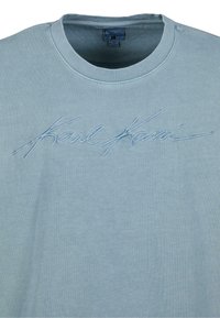 Gaiši zils kokvilnas džemperis ar ribotu apkakli. Priekšpusē ir izšūts "Karl Kani" logo, kas ir atbilstošā krāsā.