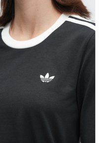 Detailný záber na osobu s čiernym tričkom Adidas s bielym golierom, pruhmi na pleciach a logom trefoil na hrudi.