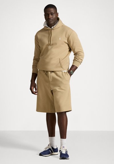 Beige pullover hoodie met een voorzak en een capuchon met trekkoord, in combinatie met beige shorts. Draagt met marineblauwe sneakers en witte crew sokken.