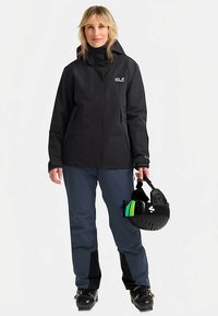 Zwarte waterdichte jas met hoge hals, gecombineerd met blauwe skibroek. De persoon houdt een zwarte helm met groene accenten vast.