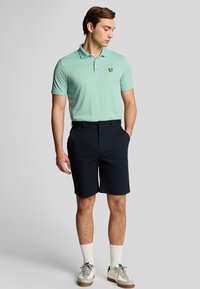 Lys grøn polo skjorte med et lille gult logo, parret med mørk marineblå shorts. Fuldført med hvide strømper og grå sneakers.