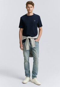 Tmavomodré bavlnené tričko, svetlomodré džínsy, so sivým sveterom zviazaným v páse. Model stojí v bielych teniskách na neutrálnej pozadí.