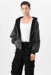 Schwarze, kurze Kapuzenjacke mit Reißverschluss und großer Kapuze, kombiniert mit strukturierten schwarzen Hosen mit fransigen Akzenten. Darunter ein weißes Tanktop.