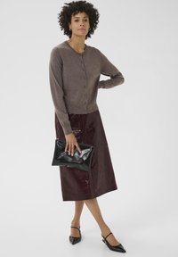 Brown knitted cardigan with buttons, paired with a burgundy glossy skirt, black clutch, and black pointed-toe shoes.