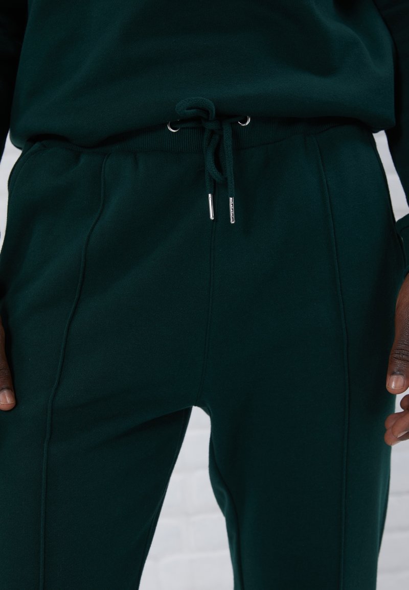 Pantaloni morbidi di colore verde scuro realizzati in materiale morbido, dotati di un elastico in vita con estremità metalliche e linee cucite lungo il lato per un tocco di dettaglio in più.