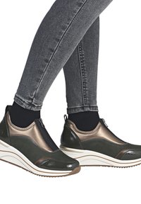 Grüne und bronzefarbene Sneaker mit einem Reißverschluss, glatter Textur und dicker weißer Zwischensohle, kombiniert mit dunklen Jeans.