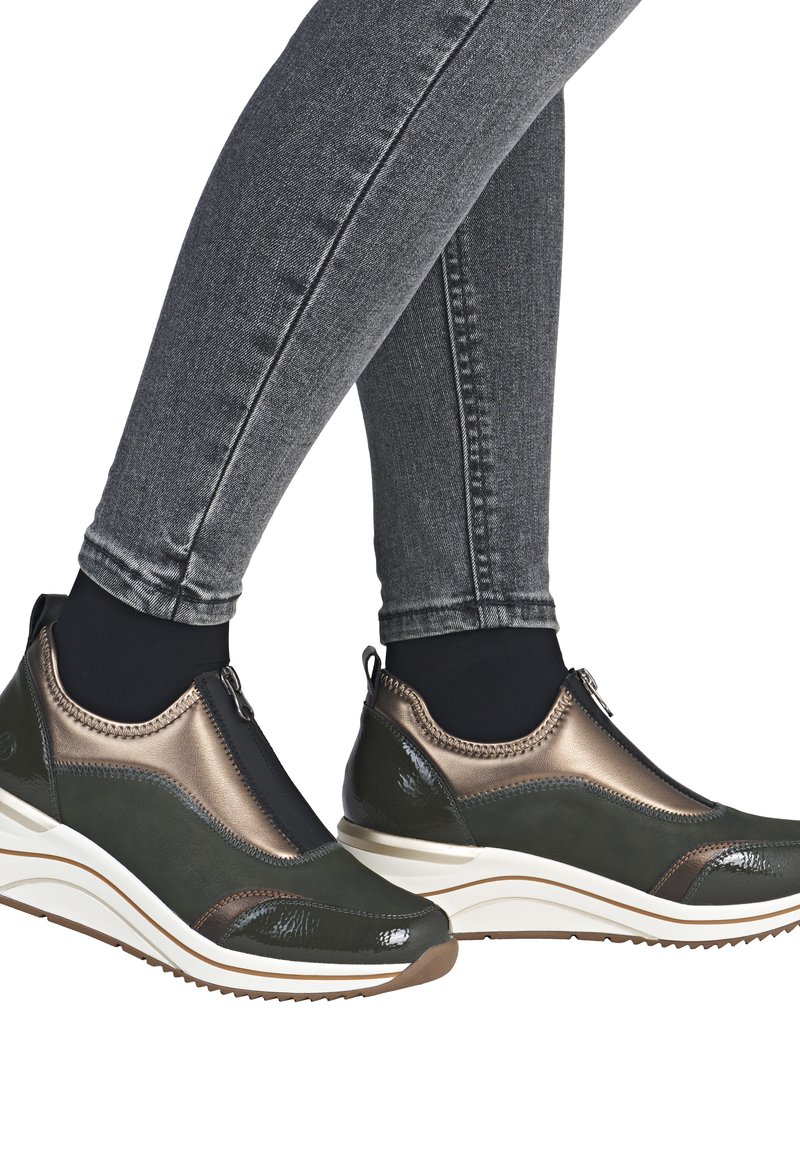 Grüne und bronzefarbene Sneaker mit einem Reißverschluss, glatter Textur und dicker weißer Zwischensohle, kombiniert mit dunklen Jeans.