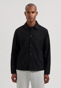 Svart teksturert skjortejakke med mønstret overflate, med krage og knappestenging, brukt over en svart skjorte.