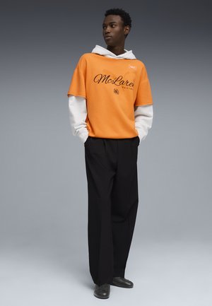 Ung mand iført en orange McLaren Racing kortærmet sweatshirt over en hvid langærmet hættetrøje, sorte bukser og sorte sko, stående med hænderne i lommerne.