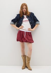 Veste en denim avec manches retroussées, t-shirt blanc avec impression rouge, jupe bordeaux avec cordon de serrage, et bottes de cowboy en daim beige.