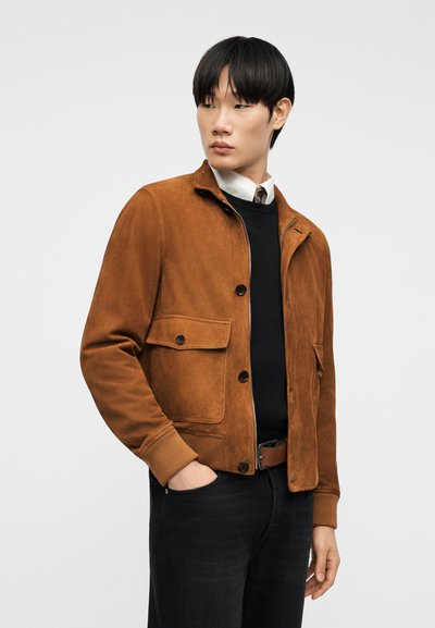 Young man wearing a brown suede jacket over a black sweater, white shirt, and black jeans, with one hand in pocket, looking to the side.