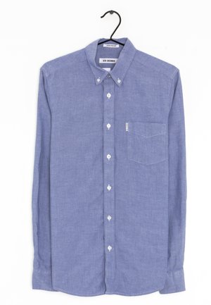 Camisa de manga larga color azul claro con bolsillo en el pecho, cuello abotonado y botones blancos, colgada en una percha negra.