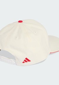 Gorra de béisbol color crema con visera curvada, complementada con detalles rojos y bordados, con agujeros de ventilación y una correa ajustable.