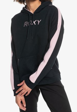 Personne portant un sweat à capuche noir avec des rayures roses le long des manches et le logo "ROXY" sur la poitrine, associé à un pantalon noir.