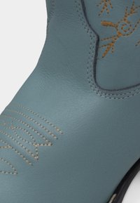 Κοντινό πλάνο γαλάζινης δερμάτινης μπότας με διακοσμητική ταμπά ραφή στο μπροστινό μέρος και στο επάνω μέρος της μπότας.