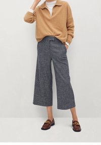 Sweter w jasnobrązowym kolorze, o miękkiej teksturze, z kołnierzem i luźnym krojem. Szerokie spodnie w szare kratki. Brązowe loafersy z akcentami złotego hardware'u.
