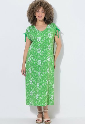 Kobieta o kręconych włosach, ubrana w zieloną sukienkę midi z białymi abstrakcyjnymi wzorami i związanymi rękawami, w beżowych sandałach, stojąca przy białej ścianie.
