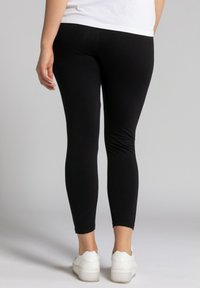 Leggings noirs en matière extensible, design ajusté, longueur cheville, associés à des baskets blanches, vus de derrière.